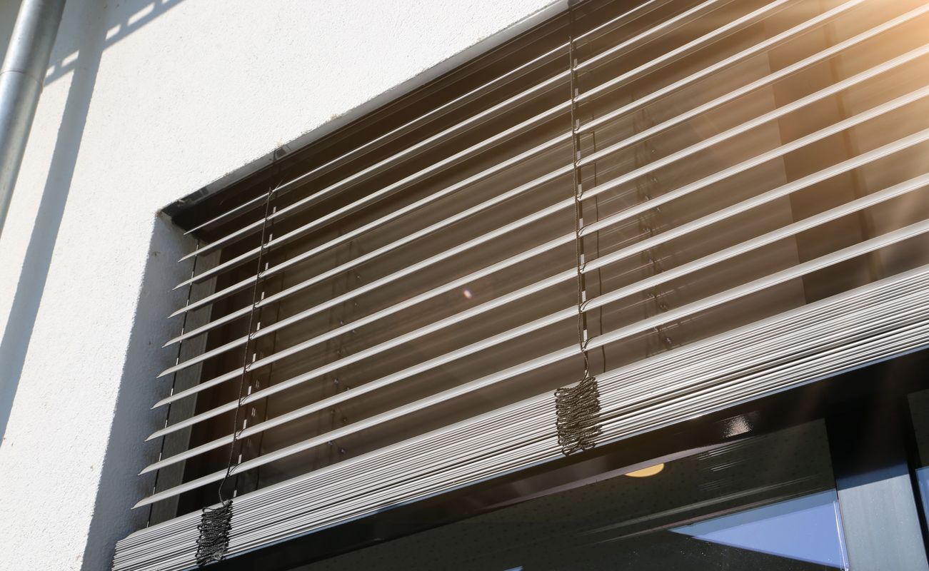 Fenster mit horizontalen Jalousien, die teilweise geöffnet sind, und einem hellen Lichtschein im oberen rechten Bereich.